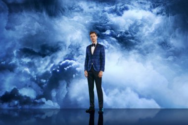 Mavi desenli smokin ve papyonlu şık bir adam dramatik bulutlu gökyüzü önünde güven içinde poz veriyor, erkek modasını, zarafetini, yaşam tarzını ve portre fotoğrafçılığını simgeliyor..