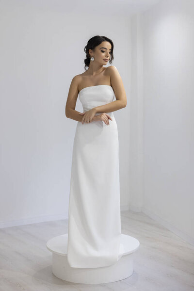 Шикарная модель в простом белом платье без бретелек позирует в яркой студии