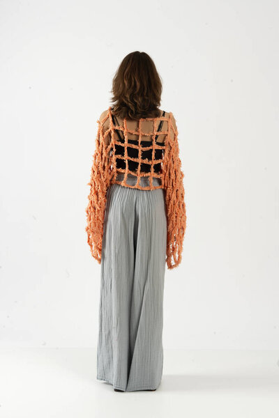 Вид сзади на женщину в оранжевом вязаном свитере поверх черной кукурузы с распущенными серыми брюками.