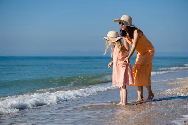 Mother and daughter in summer dresses and hats play at sea beach, walking barefoot and smiling, family vacation