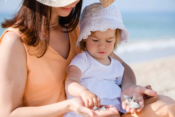 Anne ve bebek bebek portresi yaz güneşli sahilde deniz kabuklarıyla oynuyor, şapka takıyor.