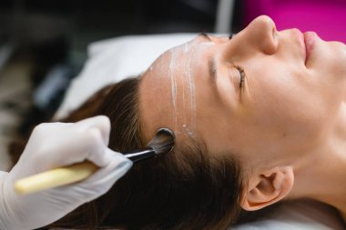 Cosmetologist apply white cream peeling mask on female face with brush in professional beauty salon, skin care treatment