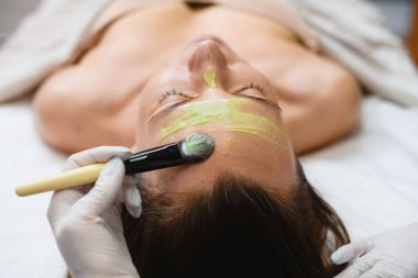 Cosmetologyst apply green face peeling anti-aging mask with brush, rejuvenating treatment and skin care