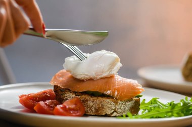 Close-up of a poached egg sandwich on a white plate. The girl is holding a fork and a knife, cutting a poached egg.