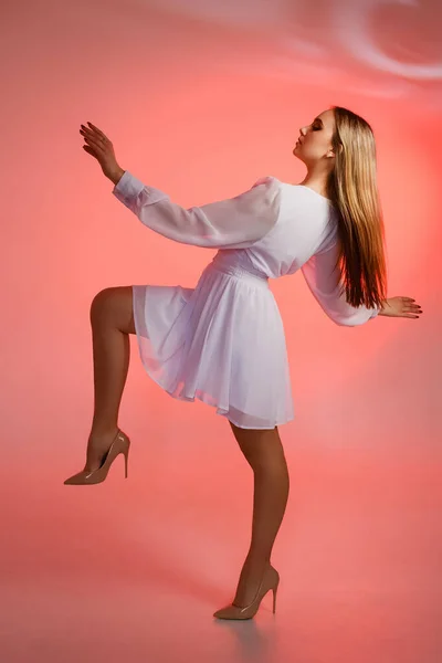 Beautiful girl in a white dress and high-heeled shoes on a pink background. The girl stands with her leg and arms raised, the dress flutters in the wind.