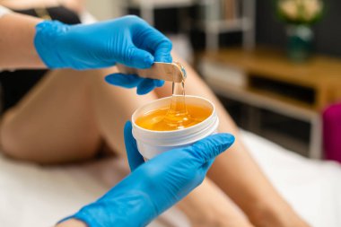 Ahşap bir çubukta karıştırmak için sıvı sarı macun, yakın plan. Eldivenli kadın elleri bir kavanoz tüy dökülme macunu tutuyor..