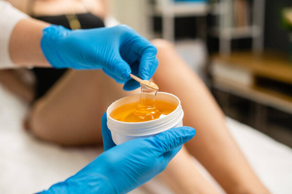 Жидкая желтая паста для укладки на деревянную палку крупным планом. Женские руки в перчатках держат банку пасты для депиляции волос.