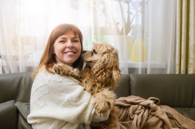 Genç bir kadın, odada kanepede otururken Amerikalı bir Cocker Spaniel 'i kollarında tutuyor. Pencereden gelen ışık.