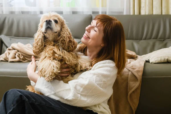 Genç bir kadın, odada koltuğun yanında otururken Amerikalı bir Cocker Spaniel 'i kollarında tutuyor. Pencereden gelen ışık.