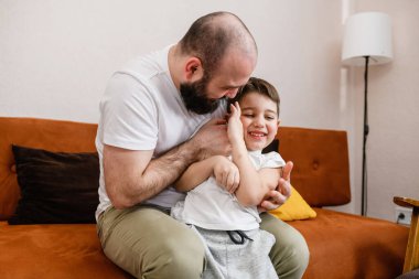 Baba küçük oğlunu rahat koltukta gıdıklıyor, aile içeride oynuyor, sevgili anaokulu bebeği, sevimli babalık