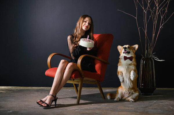 A teenage girl sits in a red chair holding a birthday cake. The corgi dog wears a red bow tie and sits next to the chair. The dog sits on its hind legs.