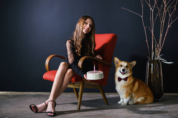 A teenage girl sits in a red chair holding a birthday cake. The corgi dog wears a red bow tie and sits next to the chair.