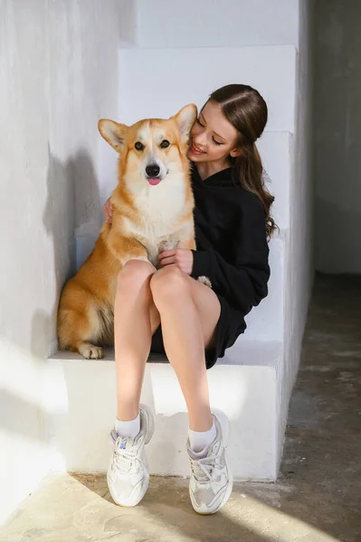 一个少女和她的科尔吉犬坐在一起 这个女孩穿着一件黑色运动衫和短裤 一个女孩和一只狗坐在屋里的白色台阶上 — 图库照片