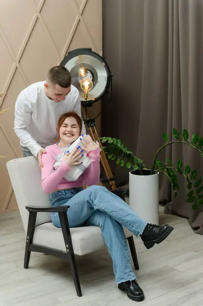 A man gives a gift to a girl. A man hugs a girl from behind. The girl is sitting in a chair.