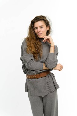 Beyaz arka planda çekici bir genç kadın. Ellerin yanağında olsun. Kız gri modern bir kazak ve geniş gri bir pantolon giyiyor. Uzun dalgalı saçlar..