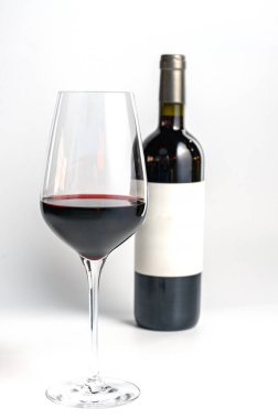 Arka planda bulanık bir şarap şişesi olan temiz beyaz bir zemin üzerine odaklanmış bir kadeh kırmızı şarap. Şişenin etiketi boş, markalaşma ya da ürün modellemeleri için mükemmel..