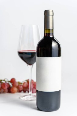 Beyaz arka plan odaklı kırmızı şarap şişesi, bir bardak kırmızı şarap ve arka planda hafifçe bulanık üzümler. Şarap ve gurme projeleri için rafine bir kompozisyon..