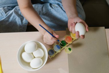 Paskalya yumurtasını yeşil boyayla boyayan birinin elleri. Masanın üzerinde bir fırça, içinde boya olan bir palet ve beyaz yumurtalı bir kase var..