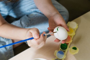 Paskalya yumurtasını yeşil boyayla boyayan birinin elleri. Masanın üzerinde bir fırça, içinde boya olan bir palet ve beyaz yumurtalı bir kase var..