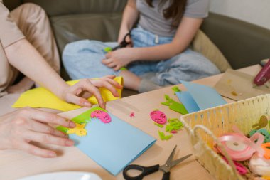 Bir anne ve kızın renkli keçeli kalemlerle Paskalya tebrik kartları hazırlaması. Masaya makas, kurdele ve el işi gereçleri serildi. Kapalı alan ayarları.