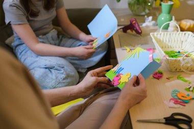 Bir anne ve kız, mavi kart, keçeli kalem ve kurdele kullanarak Paskalya tebrik kartları hazırlıyorlar. Masa renkli dekorasyon, makas ve tutkal tabancasıyla dolu..