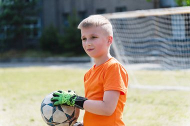Kaleci çocuk topu kalenin önünde tutuyor. Turuncu tişörtlü ve elinde futbol topu tutan yeşil eldivenli genç, kalenin önünde duruyor..