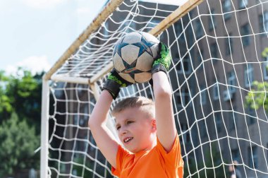 Kaleci çocuk kafasının üstünde futbol topu yakalıyor. Turuncu tişörtlü ve yeşil eldivenli genç gol önünde topu kafasının üstünde yakalıyor. Yukarı açı, arkaplanda ağ.