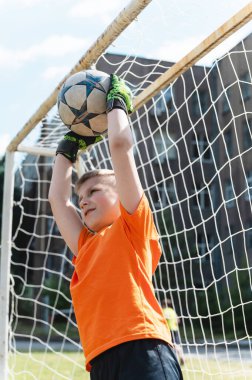 Kaleci çocuk kafasının üstünde futbol topu yakalıyor. Turuncu tişörtlü ve yeşil eldivenli genç gol önünde topu kafasının üstünde yakalıyor. Yukarı açı, arkaplanda ağ.
