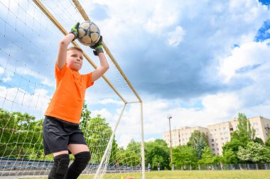 Turuncu tişörtlü ve yeşil eldivenli bir çocuk kurtarış sırasında futbol topunu kafasının üstünde tutuyor. Geniş açı görünümü, kale direği ve arkaplanda bulutlu gökyüzü.