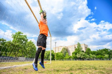 Turuncu tişörtlü ve yeşil eldivenli çocuk kalenin önündeki futbol topunu yakalamak için zıplıyor. Geniş açı, çimenlik alan, arka planda dramatik gökyüzü.