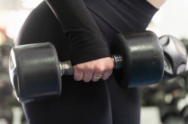 Spor salonunda spor yaparken büyük siyah bir dambılı tutan bir kadının yakın çekimi. Kadın siyah tayt ve uzun kollu bir bluz giyiyor. Hafif ışıklandırma, arka planda bulanık spor aletleri..