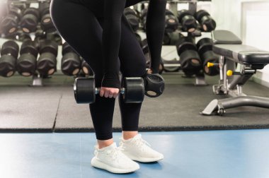 Spor salonunda siyah tayt ve beyaz spor ayakkabılı bir kadının dambıl tutuşu. O, kaldırma pozisyonunda öne eğildi. Kapalı alanda ışıklandırma, arka planda spor aletleri..
