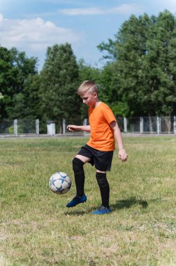 Turuncu tişörtlü bir çocuk güneşli bir çimenlikte futbol topuyla tekme atıyor. Hokkabazlık yaparken konsantrasyonunu kaybediyor. Parlak gün ışığı ve yeşil arkaplan.