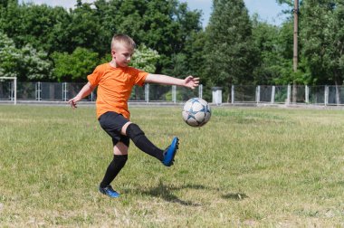 Turuncu tişörtlü ve siyah spor giysili bir çocuk çimenli bir alanda futbol topunu havada tekmeliyor. Parlak gün ışığında yeşil ağaçlar ve arka planda çitlerle eylem anı.