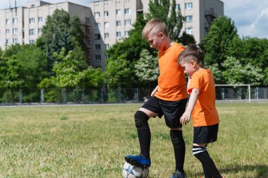 Turuncu tişörtlü iki genç çimenli bir sahada futbol topu sahibi olmak için kavga ediyor. Bir çocuk topa basarken diğeri topu almaya çalışıyor. Yeşil ağaçlar ve binalarla aydınlık bir gün..