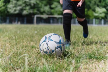 Siyah şortlu ve çoraplı çocuğun bacaklarının çimenli sahada futbol topunu tekmelemek üzere olduğu yakın plan. Güneşli bir gün, kale direği ve arka planda ağaçlar, aksiyon anı.
