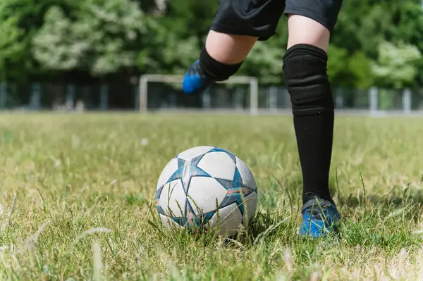 Siyah şortlu ve çoraplı çocuğun bacaklarının çimenli sahada futbol topunu tekmelemek üzere olduğu yakın plan. Güneşli bir gün, kale direği ve arka planda ağaçlar, aksiyon anı.