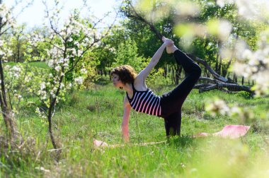 Yeşil tarlada pembe hasırda şeker kamışı pozu veren kadın, tek ayak üzerinde dengede duruyor, arka ayağını eliyle tutuyor, çizgili üst, çiçek açan ağaçlar..
