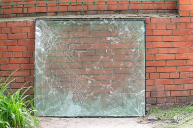 Kırmızı tuğla bir duvara dayanan örümcek ağı kırıklarının olduğu büyük çatlak bir cam panel. Gün ışığında dışarıda çekilmiş. Etrafında yeşillik var..