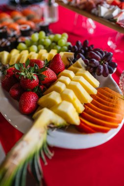 Kırmızı bir masa örtüsü üzerinde dilimlenmiş ananas, papaya, çilek ve üzümle beyaz bir tabağın yakın çekimi. Açık büfe veya kutlama için renkli tropikal meyve aranjmanı.