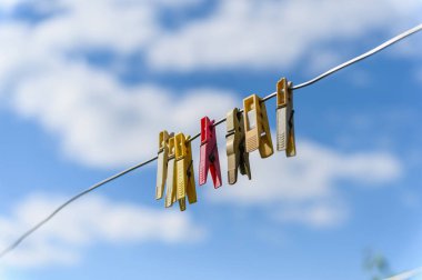 Renkli plastik mandallar beyaz bulutlu açık mavi gökyüzüne karşı bir tel üzerinde asılı. Çapraz kompozisyon, açık hava sahnesi, güçlü güneş ışığı ve yumuşak kontrast.