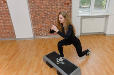 Siyah spor giysili genç bir kadın gri bir aerobik platformunda tek ayakla hamle yapıyor. Ahşap zemini ve tuğla duvarı olan parlak bir spor salonu..