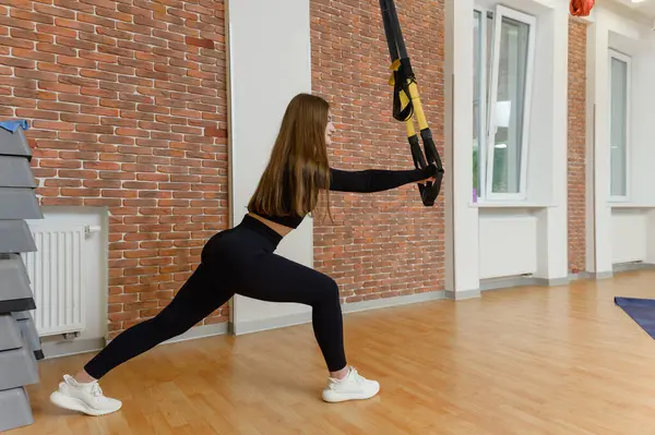 Spor kıyafetli genç bir kadın spor salonunda trx fitness kayışları kullanarak esneme hareketleri yapıyor. Kollarını açıp öne doğru eğiliyor..