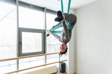 Atletik kadın tersten poz veriyor. Gökyüzü ipekleri, bacakları açık ve vücudu zarif bir şekilde geniş pencereli, modern bir stüdyoda baş aşağı asılı..