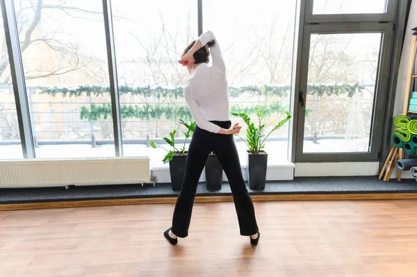Beyaz bluzlu ve siyah pantolonlu bir kadın geniş bacaklı bir poz vermiş, kolları arkadan bükülmüş, büyük pencereleri ve bitkileri olan parlak bir stüdyoda dans esneme hareketleri yapıyor..