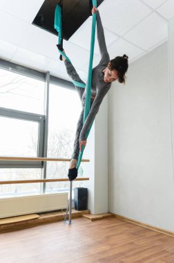 Gri elbiseli kadın, parlak bir dans stüdyosunda ya da ahşap zemin ve geniş pencereli yoga stüdyosunda uzun bacaklı bir poz vererek deniz mavisi kumaşı kullanarak zarif bir şekilde hava esneme hareketi yapıyor..