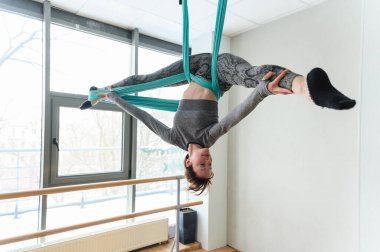 Atletik kadın tersten poz veriyor. Gökyüzü ipekleri, bacakları açık ve vücudu zarif bir şekilde geniş pencereli, modern bir stüdyoda baş aşağı asılı..