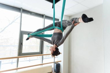 Atletik kadın tersten poz veriyor. Gökyüzü ipekleri, bacakları açık ve vücudu zarif bir şekilde geniş pencereli, modern bir stüdyoda baş aşağı asılı..