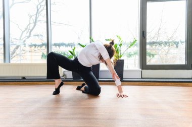 Siyah pantolonlu ve beyaz bluzlu kadın, uzun pencereler ve kapalı bitkilerin yanındaki ahşap bir zeminde, kıvrımlı duruşu ve genişleyen koluyla, düşük dans pozunda zemin esnetiyor..