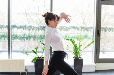Beyaz bluzlu ve siyah pantolonlu kadın kıvrımlı kolları ve bükülmüş duruşu ile dinamik dans hareketleriyle büyük pencereleri ve yeşil bitkileri olan bir stüdyoda zarifçe hareket ediyor..